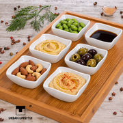 Sechs weiße eckige Nami-Schälchen mit Hummus, Sojasauce, Nüssen, Oliven und Edamame auf einem Holztablett